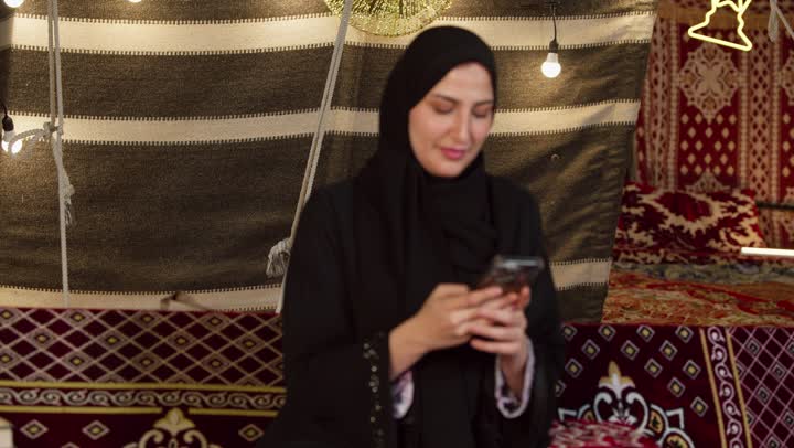 استخدام وسائل التواصل الاجتماعي عبر الجهاز التقني الحديث, كشتة بر وتخييم في صحراء الامارات، ديكور وزينة شهر رمضان المبارك، تصوير مقرب لسيدة عربية خليجية إماراتية محجبة ترتدي عباءة تقوم باستخدام الهاتف المحمول بإيماءات السعادة والسرور
