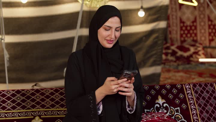 كشتة بر وتخييم في صحراء الامارات، ديكور وزينة شهر رمضان المبارك، التواصل مع الأهل والاصدقاء عبر الجهاز التقني الحديث, تصوير مقرب لسيدة عربية خليجية إماراتية محجبة ترتدي عباءة تقوم باستخدام الهاتف المحمول بإيماءات السعادة والسرور