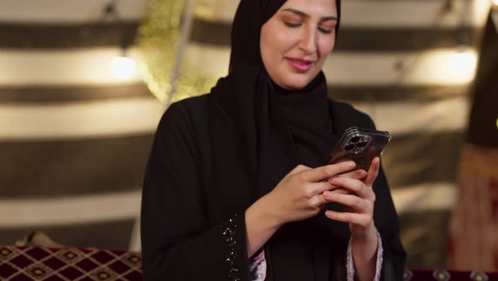 كشتة بر وتخييم في صحراء الامارات، ديكور وزينة شهر رمضان المبارك، التواصل مع الأهل والاصدقاء عبر الجهاز التقني الحديث, تصوير مقرب لسيدة عربية خليجية إماراتية محجبة ترتدي عباءة تقوم باستخدام الهاتف المحمول بإيماءات السعادة والسرور