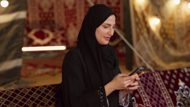 استخدام وسائل التواصل الاجتماعي عبر الجهاز التقني الحديث, كشتة بر وتخييم في صحراء الامارات، ديكور وزينة شهر رمضان المبارك، تصوير مقرب لسيدة عربية خليجية إماراتية محجبة ترتدي عباءة تقوم باستخدام الهاتف المحمول بإيماءات السعادة والسرور