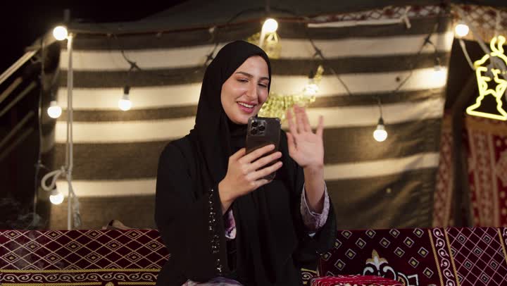 Ramadan decorations and adornments, communicating with family and friends via modern technology, a close-up of a veiled Emirati Gulf Arab woman wearing a black abaya making a video call using a mobile phone, a desert outing and camping in the UAE desert, a quiet night atmosphere in a traditional tent