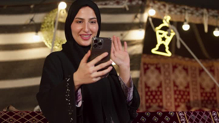 Ramadan decorations and adornments, communicating with family and friends via modern technology, a close-up of a veiled Emirati Gulf Arab woman wearing a black abaya making a video call using a mobile phone, a desert outing and camping in the UAE desert, a quiet night atmosphere in a traditional tent