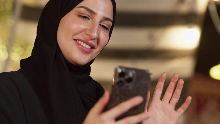 Greetings and peace, communicating with family and friends via modern technology, a close-up of a veiled Emirati Gulf Arab woman wearing an abaya making a video call using a mobile phone with happy expressions, a desert outing and camping in the UAE desert, a quiet night atmosphere in a traditional tent
