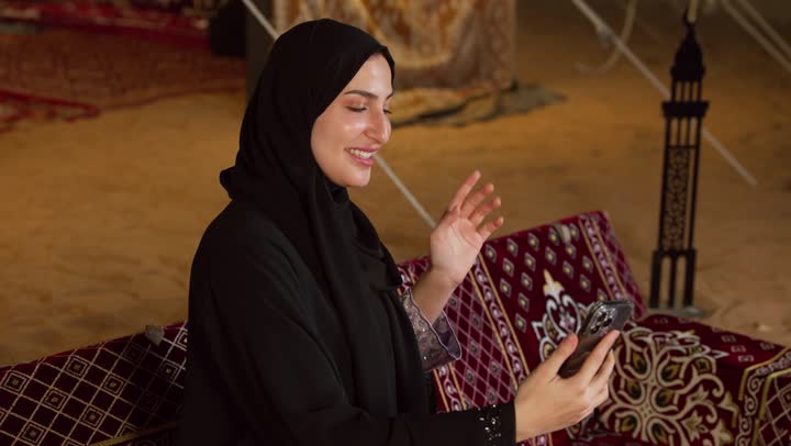 Greetings and peace, communicating with family and friends via modern technology, a close-up of a veiled Emirati Gulf Arab woman wearing an abaya making a video call using a mobile phone with happy expressions, a desert outing and camping in the UAE desert, a quiet night atmosphere in a traditional tent