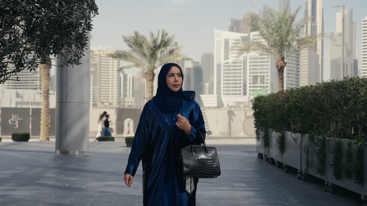 Attention to elegance and outward appearance, the concept of femininity and softness, a smiling Saudi Arabian Gulf woman wearing an abaya and hijab looking at something with gestures of pleasure and self-confidence, an Emirati woman wearing a colorful abaya and a black bag walking with confident steps