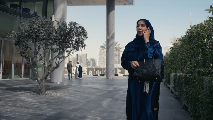 امرأه عربية خليجية سعودية تقوم بإحتضان صديقتها بإيماءات الفرح والابتسامه، سيده سعوديه ترتدي العباءه والحجاب تجري مكالمه الهاتف المحول، ايماءات الاحتضان والمحبة, قضاء أوقات ممتعة مع الأصدقاء، استخدام التقنيات الحديثه