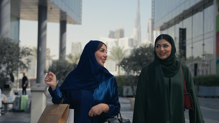 Benefiting from Black Friday discounts, the joy of sharing with friends in choosing things and supplies from shopping malls, an Emirati woman walking with her friend, the positive impact of shopping on women, two smiling Saudi Arabian Gulf Arab friends carrying paper bags