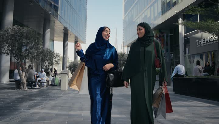 Benefiting from Black Friday discounts, the joy of sharing with friends in choosing things and supplies from shopping malls, an Emirati woman walking with her friend, the positive impact of shopping on women, two smiling Saudi Arabian Gulf Arab friends carrying paper bags