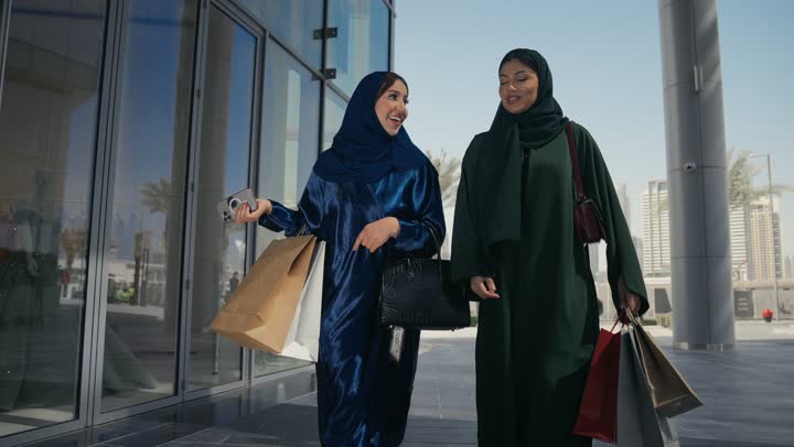 The joy of sharing gifts with friends from shops and malls on White Friday. Two Emirati Gulf Arab friends carry paper bags and shop in the mall. Buying from luxury Emirati brands. The positive impact of shopping on women.