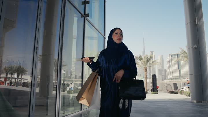 امراه عربيه خليجيه اماراتيه ترتدي عباءه ملونه تقوم بالنظر الي شئ ما، سيده سعوديه تسير فى احد الاماكن الخارجيه وتقوم بحمل كيس ورقي، التسوق وشراء الهدايا والمستلزمات، التمعن في شئ ما