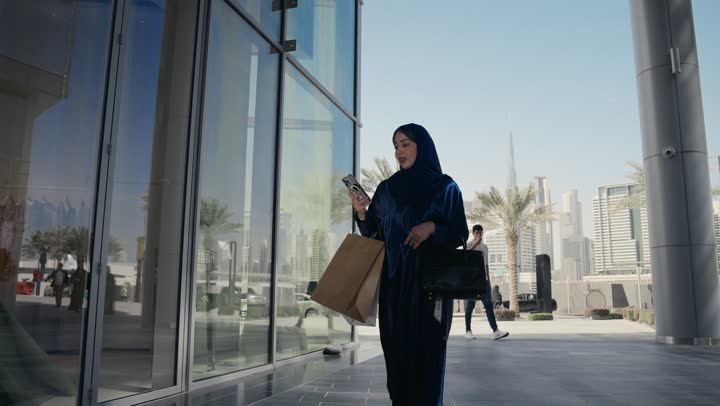 Enjoying shopping in malls and commercial centers, an Emirati woman holding a mobile phone, taking advantage of offers and discounts, a Saudi Arabian Gulf woman wearing an abaya holding paper shopping bags, using modern devices and technologies