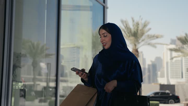 الاستمتاع بالتسوق في المولات والمراكز التجارية،  سيده اماراتيه تحمل بيدها الهاتف المحمول، الاستفادة من العروض والخصومات، امرأة عربية خليجية سعودية ترتدي العباءة تحمل بيدها اكياس التسوق الورقيه، استخدام الاجهزة والتقنيات الحديثه