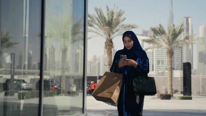 الشراء عبر الاونلاين، عروض وتخفيضات الجمعة البيضاء، ايماءات وجه تدل على الإستمتاع بالتسوق و شراء الهدايا، إمرأة عربية سعودية خليجية ترتدي العباءه والحجاب تحمل بيدها أكياس، التسوق الإلكتروني الآمن، سيده اماراتيه تسير في احد الاماكن الخارجيه