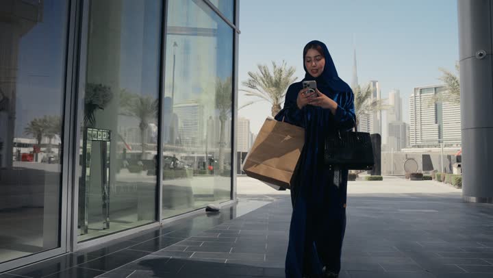 الشراء عبر الاونلاين، عروض وتخفيضات الجمعة البيضاء، ايماءات وجه تدل على الإستمتاع بالتسوق و شراء الهدايا، إمرأة عربية سعودية خليجية ترتدي العباءه والحجاب تحمل بيدها أكياس، التسوق الإلكتروني الآمن، سيده اماراتيه تسير في احد الاماكن الخارجيه
