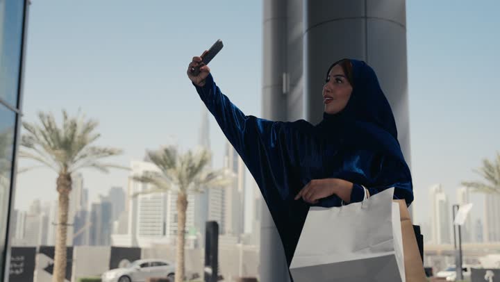 A Saudi Arabian Gulf woman is making a video call with someone, a Saudi lady wearing an abaya and hijab carrying colorful paper bags, shopping and buying gifts, enjoying shopping time, using modern technology