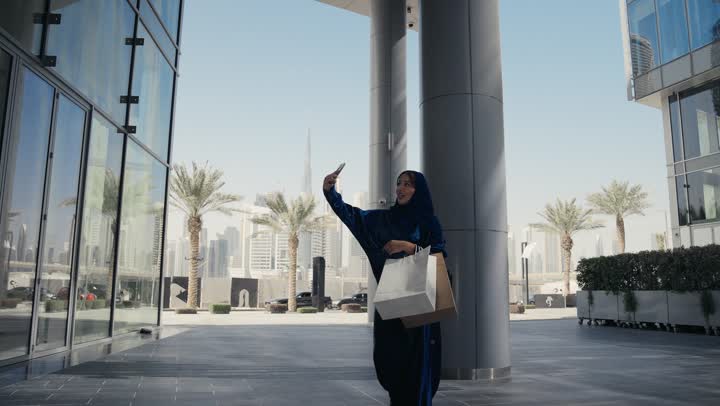 امرأه عربية خليجية سعودية تقوم باجراء مكالمه فيديو مع احد ما، سيده سعوديه ترتدي العباءه والحجاب تحمل اكياس ورقيه ملونه، التسوق وشراء الهدايا, الاستمتاع بوقت التسوق، استخدام التقنيات الحديثه