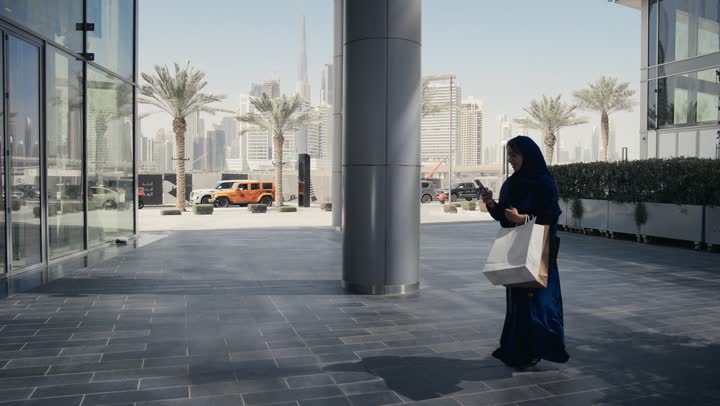 ايماءه الاعجاب والاندهاش اثناء التسوق، امرأه عربيه خليجيه اماراتيه ترتدي عباءه رماديه تسير في احد اماكن التسوق، سيده سعوديه تحمل اكياس ورقيه ملونه تلتقط صوره لاحدي المنتجات العصريه، اماكن التسوق في الامارات العربيه المتحده، الاهتمام بالازياء