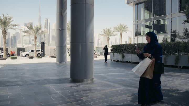 A gesture of admiration and amazement while shopping, an Arab Gulf Emirati woman wearing a gray abaya walking in a shopping area, a Saudi woman carrying colorful paper bags taking a picture of a trendy product, shopping places in the United Arab Emirates, interest in fashion
