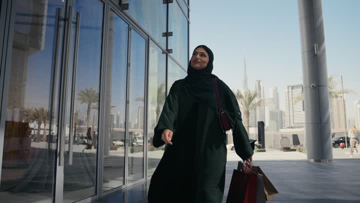 النظر والاعجاب باماكن ترفيهيه في الامارات، امرأه عربيه خليجيه اماراتيه ترتدي عباءه رماديه تسير في احد اماكن التسوق، سيده سعوديه تحمل اكياس ورقيه ملونه تنظر الي شئ ما،  مفهوم الاناقه والثقه في النفس، الاماكن السياحه في الامارات العربيه المتحده