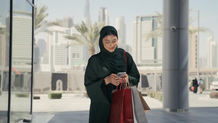 An Emirati Gulf Arab woman wearing a gray abaya walks in a shopping mall, a Saudi woman carrying colorful paper bags looks at her phone with a gesture of joy and happiness, the concept of elegance and self-confidence, tourism and entertainment in the United Arab Emirates