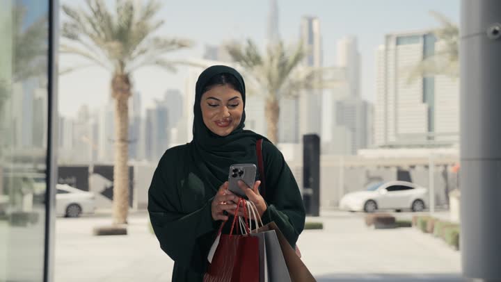 التسوق وشراء المستلزمات والهدايا, الاثر الايجابي للتسوق على نفسية النساء، امرأة عربية خليجية اماراتيه ترتدي الحجاب والعباءة السوداء تحمل بيدها أكياس ورقيه، التجول في المولات والمراكز التجارية، امرأه سعوديه تجري مكالمه فيديوباستخدام الهاتف المحمول