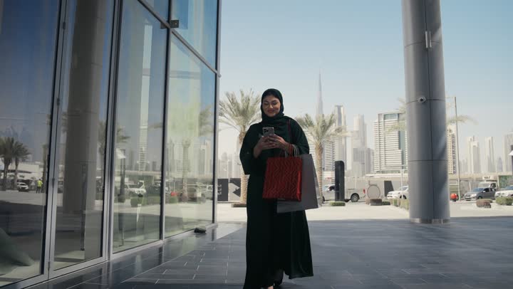 التسوق وشراء المستلزمات والهدايا, الاثر الايجابي للتسوق على نفسية النساء، امرأة عربية خليجية اماراتيه ترتدي الحجاب والعباءة السوداء تحمل بيدها أكياس ورقيه، التجول في المولات والمراكز التجارية، امرأه سعوديه تجري مكالمه فيديوباستخدام الهاتف المحمول