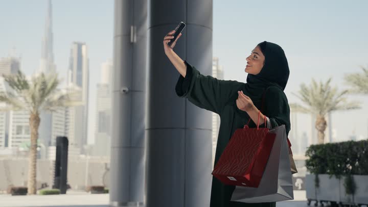 Buying some supplies and gifts, taking advantage of seasonal discounts, a veiled Saudi Arabian Gulf woman wearing a black abaya carrying colorful shopping bags, enjoying shopping and getting offers, an Emirati woman taking souvenir photos with her phone while shopping
