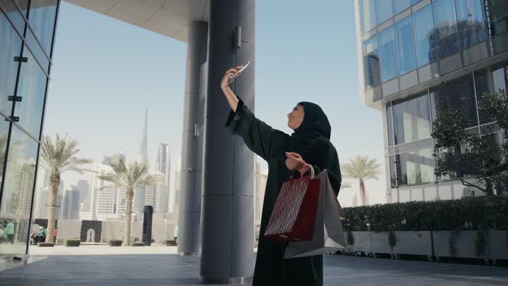 Buying some supplies and gifts, taking advantage of seasonal discounts, a veiled Saudi Arabian Gulf woman wearing a black abaya carrying colorful shopping bags, enjoying shopping and getting offers, an Emirati woman taking souvenir photos with her phone while shopping