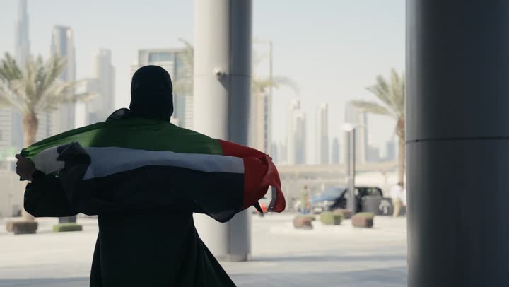 The concept of love and belonging to the country, celebrating the UAE National Day, national holidays and occasions, a portrait of an Emirati Gulf Arab woman wearing a black abaya carrying the UAE flag on her back