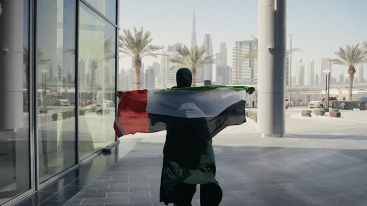 مفهوم حب الوطن والعلم، الإحتفال بالعيد الوطني الإماراتي 2 ديسمبر، إحياء ذكرى استقلال دولة الإمارات العربية المتحدة، امراة عربية خليجية إماراتية ترتدي عباءة سوداء تحمل علم الإمارات على ظهرها، الاعتزاز والفخر بإنجازات الوطن