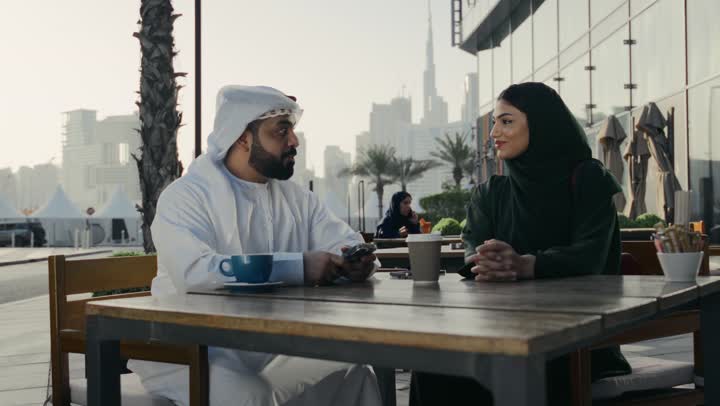 قضاء وقت ترفيهي ممتع برفقة الزوجة، تقوية روابط الألفة والمحبة بين الزوجين، رجل عربي خليجي اماراتي يرتدي الكندوره والغتره البيضاء يقوم بالتقاط صوره لزوجته في المطعم، زوجان اماراتيان يجلسان في احد المقاهي الخارجيه، تناول المشروبات الساخنه
