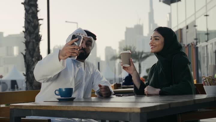 Spending a fun and entertaining time with the wife, strengthening the bonds of intimacy and love between the spouses, an Emirati Gulf Arab man wearing a white kandura and ghutra taking pictures of his wife in a restaurant, an Emirati couple sitting in an outdoor cafe, drinking hot drinks