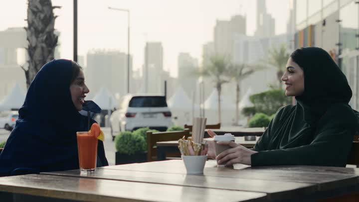 Discussions and dialogues in a calm atmosphere, two Saudi women spending pleasant times in an outdoor cafe, an Arab Gulf Emirati woman wearing a hijab and a black abaya sitting on the couch with her friend in a cafe with happy gestures, sharing cold drinks