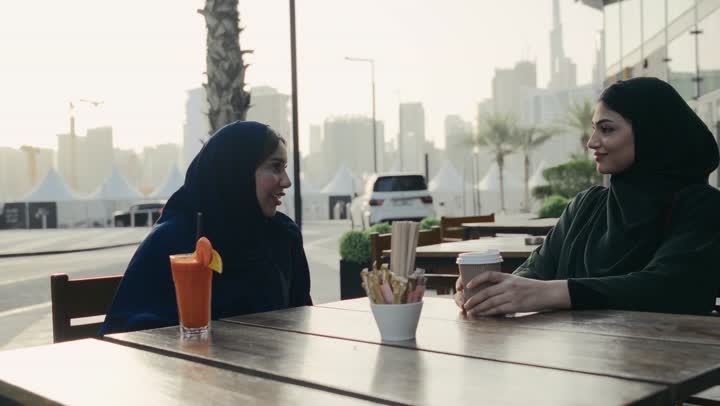 الاستمتاع بالمشروبات الباردة, النقاشات والحوارات في الأجواء الهادئة، امرأة عربية خليجية إماراتية محجبة ترتدي العباءة السوداء تجلس مع صديقتها بإحدى المقاهي, صديقتان اماراتيتان تقضيان أوقات ممتعة ومسلية، استخدام الهاتف لالتقاط صورتذكاريه