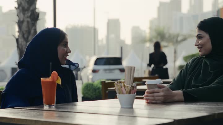 Enjoying cold drinks, discussions and dialogues in a calm atmosphere, a veiled Emirati Gulf Arab woman wearing a black abaya sitting with her friend in a cafe, two Emirati friends spending fun and entertaining times, using the phone to take souvenir photos