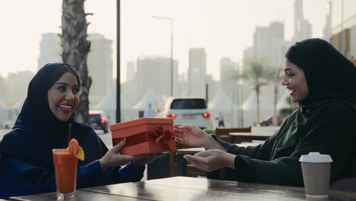 Two Emirati Gulf Arab friends enjoying chatting, enjoying refreshing drinks, friendships and getting to know each other, spending quality time, discussions and dialogues, a Saudi woman giving a gift to her friend, a gesture of surprise and pleasure
