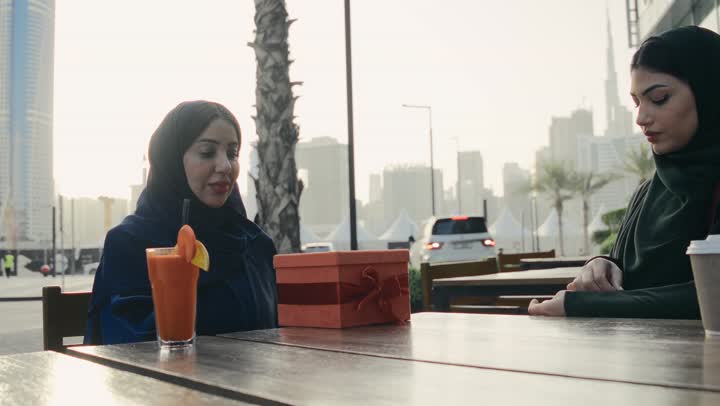 Two Saudi Arabian Gulf women sitting at a table in a cafe, two smiling Emirati friends sitting together on a pleasant outing, buying and giving gifts, friendships and getting to know each other, social relations, spending fun times with friends