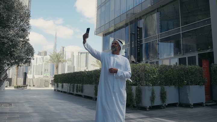 Thumbs up gestures, using a modern technology device, elegance and attention to one's outward appearance, an Emirati Gulf Arab man wearing a ghutra and kandura taking a selfie using a mobile phone in front of a company building in the Emirates, walking outdoors