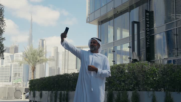 Elegance and attention to one's outward appearance, raising one's thumbs in a gesture of satisfaction and admiration, using a modern technical device, an Emirati Gulf Arab man wearing a ghutra and kandura taking a selfie using a mobile phone in front of a company building in the Emirates, walking in the open air