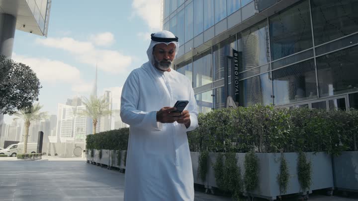استخدام جهاز تقني حديث متطور في الحياة اليومية، مفهوم الاناقة والرجولة والاهتمام بالمظهر الخارجي، رجل عربي خليجي اماراتي يرتدي الغترة والكندورة التقليدية يمسك بيده الهاتف المحمول، خلفية مبنى احدى الشركات في الامارات العربية المتحدة، التجول في الهواء الطلق