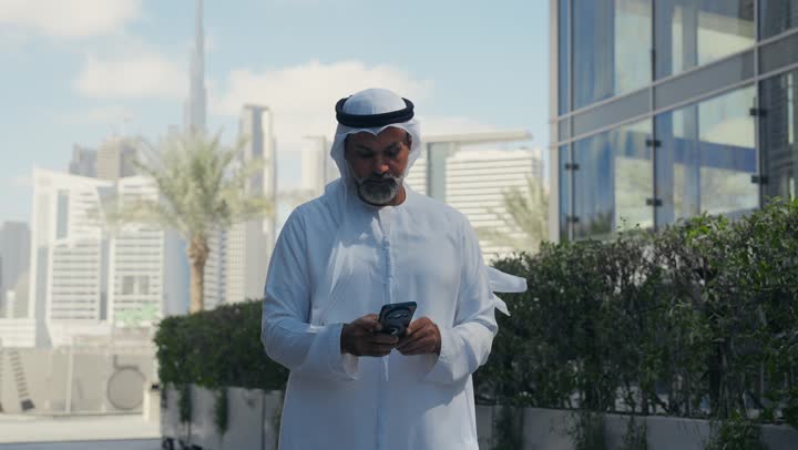 استخدام جهاز تقني حديث متطور في الحياة اليومية، مفهوم الاناقة والرجولة والاهتمام بالمظهر الخارجي، رجل عربي خليجي اماراتي يرتدي الغترة والكندورة التقليدية يمسك بيده الهاتف المحمول، خلفية مبنى احدى الشركات في الامارات العربية المتحدة، التجول في الهواء الطلق