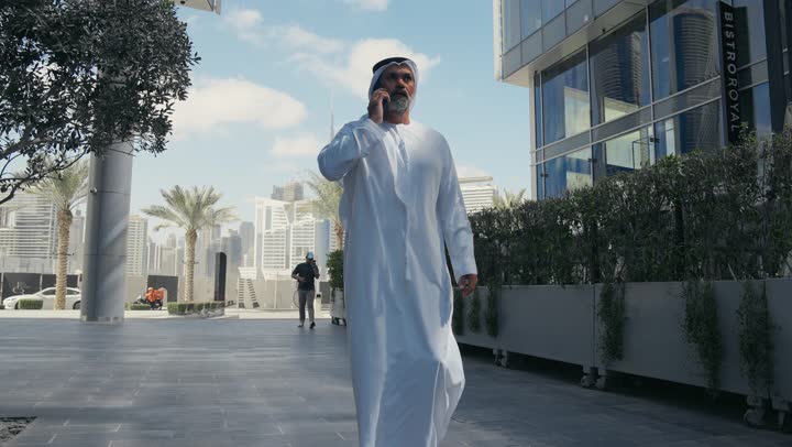 Making a phone call, using a modern, advanced technical device in daily life, elegance and attention to one's outward appearance, an Arab Gulf Emirati man wearing a traditional ghutra and kandura answering a mobile phone in front of a company building in the United Arab Emirates, walking outdoors