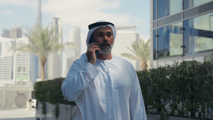 التجول في الهواء الطلق، اجراء مكالمة هاتفية، استخدام جهاز تقني حديث متطور في الحياة اليومية، الاناقة والاهتمام بالمظهر الخارجي، رجل عربي خليجي اماراتي يرتدي الغترة والكندورة التقليدية يقوم بالرد على الهاتف المحمول امام مبنى احدى الشركات في الامارات العربية المتحدة