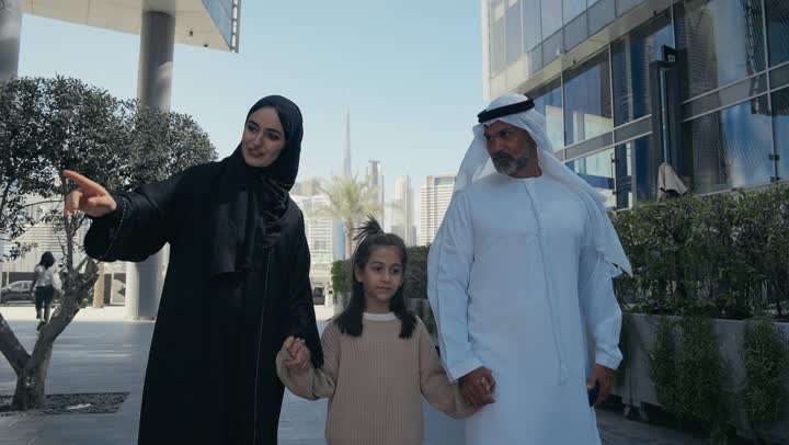 التنزه في وقت العطلة في إحدى الاماكن العامة في الإمارات, تبادل الأحاديث والأخبار بإيماءات السرور والفرح, زوجان عربيان خليجيان إماراتيان يقومان بالمشي والتجول في الهواء الطلق برفقة ابنتهم, الذكريات واللحظات العائلية السعيدة, الاشارة الى شيء ما