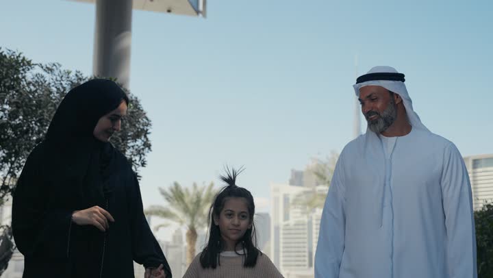 تبادل الأحاديث والأخبار بإيماءات السرور والفرح, التنزه في وقت العطلة في إحدى الاماكن العامة في الإمارات, زوجان عربيان خليجيان إماراتيان يقومان بالمشي والتجول في الهواء الطلق برفقة ابنتهم, الذكريات واللحظات العائلية السعيدة, الاشارة الى شيء ما