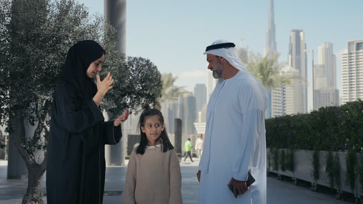 Exchanging conversations and news with gestures of pleasure and joy, taking a walk during the holiday in a public place in the Emirates, an Emirati Gulf Arab couple walking and strolling in the open air with their daughter, happy family memories and moments, pointing to something