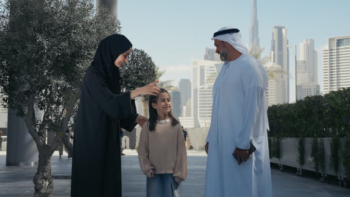 A walk during the holiday in a public place in the Emirates, exchanging conversations and news with gestures of pleasure and joy, an Emirati Gulf Arab couple walking and strolling in the open air with their daughter, happy family memories and moments, pointing to something