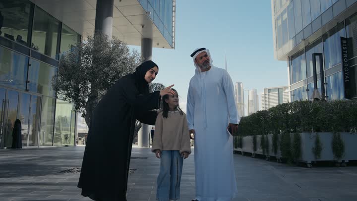 تبادل الأحاديث والأخبار بإيماءات السرور والفرح, التنزه في وقت العطلة في إحدى الاماكن العامة في الإمارات, زوجان عربيان خليجيان إماراتيان يقومان بالمشي والتجول في الهواء الطلق برفقة ابنتهم, الذكريات واللحظات العائلية السعيدة, الاشارة الى شيء ما