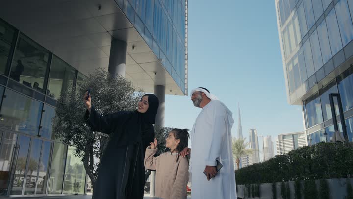 التنزه في وقت العطلة في إحدى الاماكن العامة في الإمارات, زوجان عربيان خليجيان إماراتيان يقومان بالمشي والتجول في الهواء الطلق برفقة ابنتهم, الذكريات واللحظات العائلية السعيدة, امرأة سعودية قوم بالتقاط صور السيلفي التذكارية باستخدام الهاتف المحمول بإيماءات السعادة