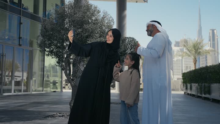 التنزه في وقت العطلة في إحدى الاماكن العامة في الإمارات, زوجان عربيان خليجيان إماراتيان يقومان بالمشي والتجول في الهواء الطلق برفقة ابنتهم, الذكريات واللحظات العائلية السعيدة, امرأة سعودية قوم بالتقاط صور السيلفي التذكارية باستخدام الهاتف المحمول بإيماءات السعادة